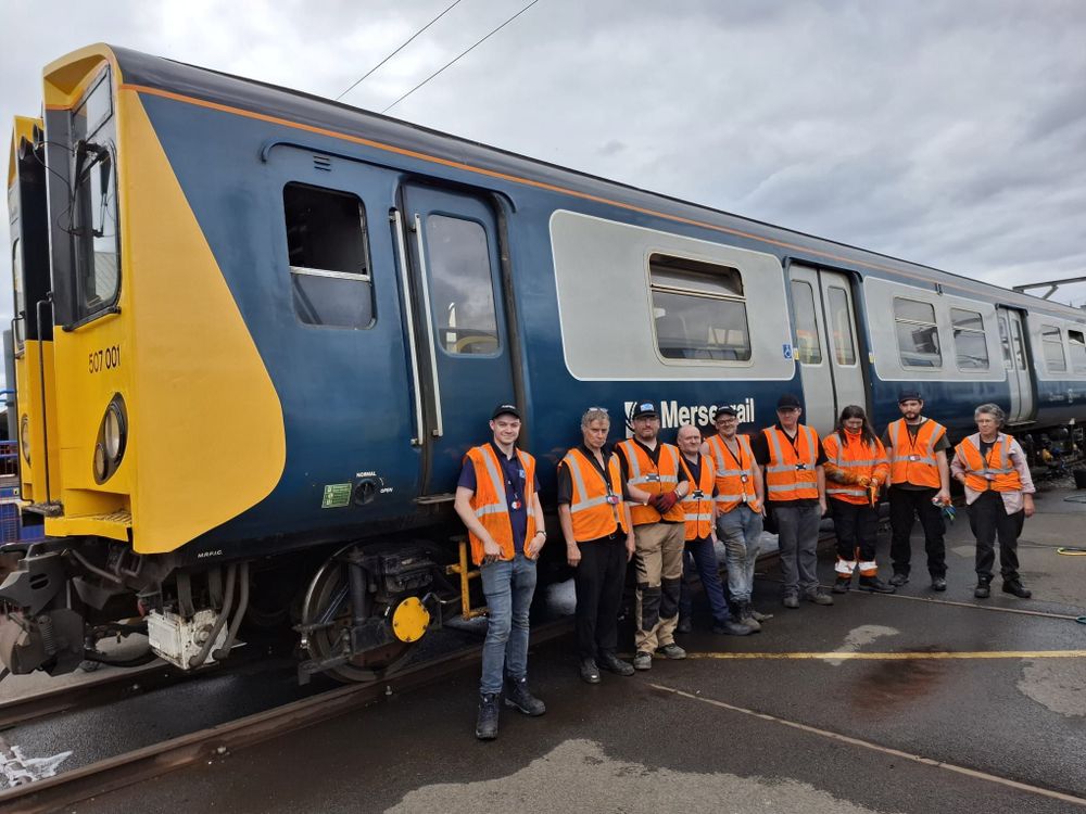 Nine people in high-vis jackets pose on front of 507001 on an outside siding at Litchurch Lane works