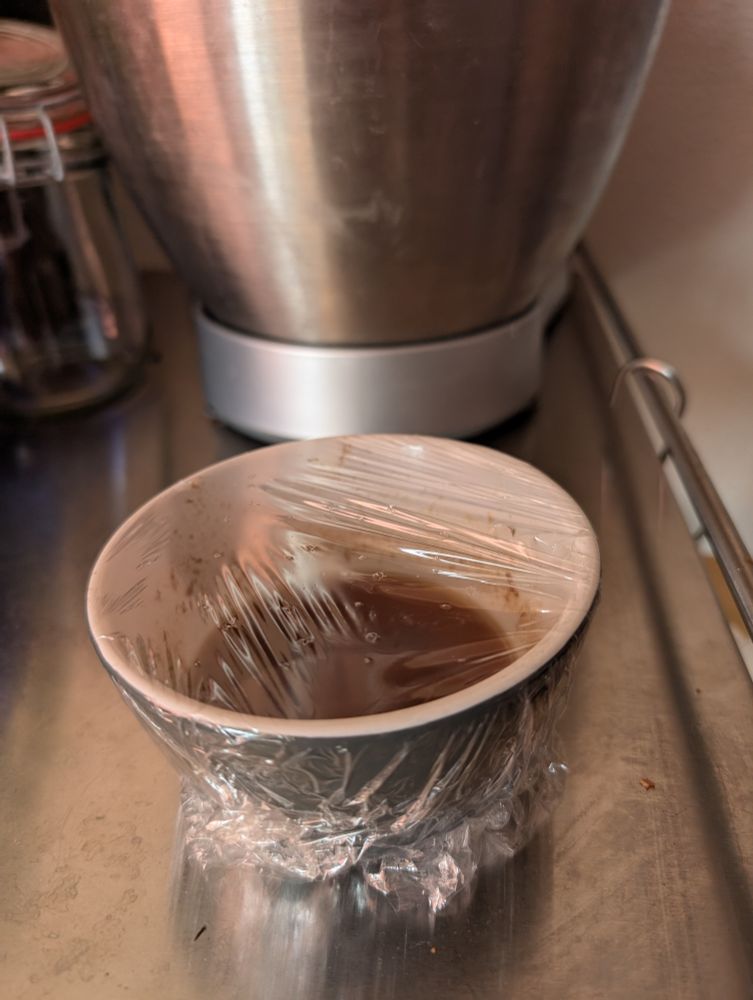 Een kom met jam en balsamico, afgedekt met huishoudfolie met gaatjes er ln om fruitvliegjes te vangen.