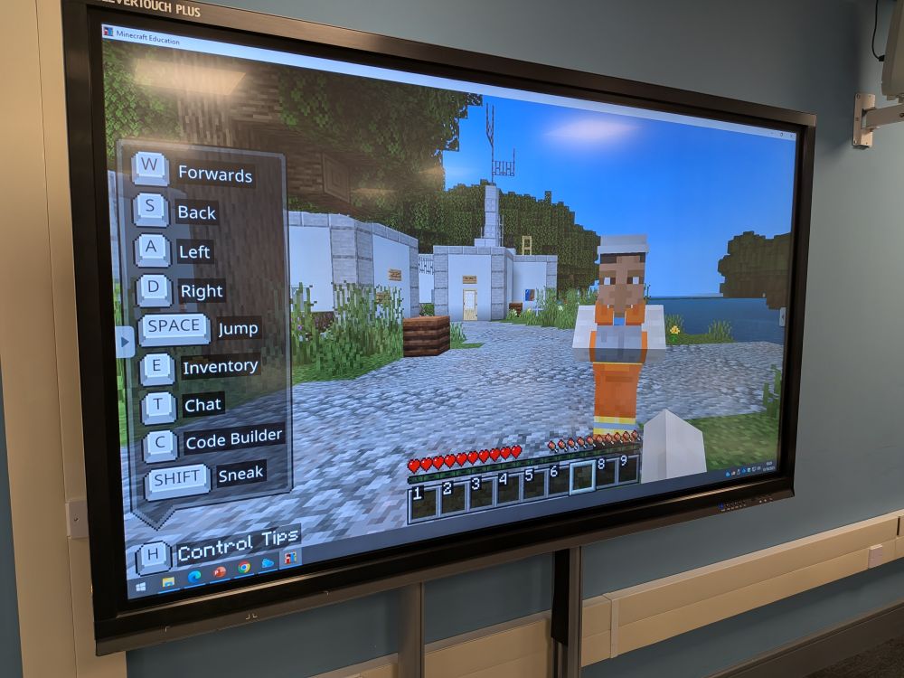 A screenshot from the Minecraft game with a river, buildings, and a person visible.