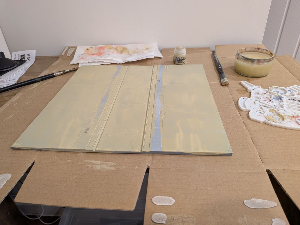 A plastic game board tile of a straight roadway being painted an off white colour, surrounded by painting equipment.