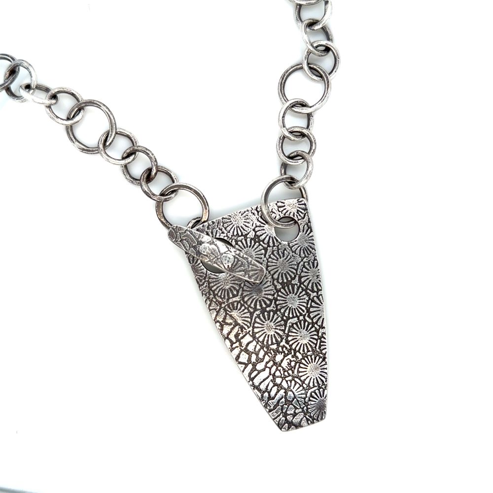 The photo is of a sterling silver necklace against a white background. The shape of the pendant is an elongated squared off triangle. It is highly textured with abstract floral patterns. The pendant attaches to a handmade heavy silver chain with a toggle clasp with the same pattern. 