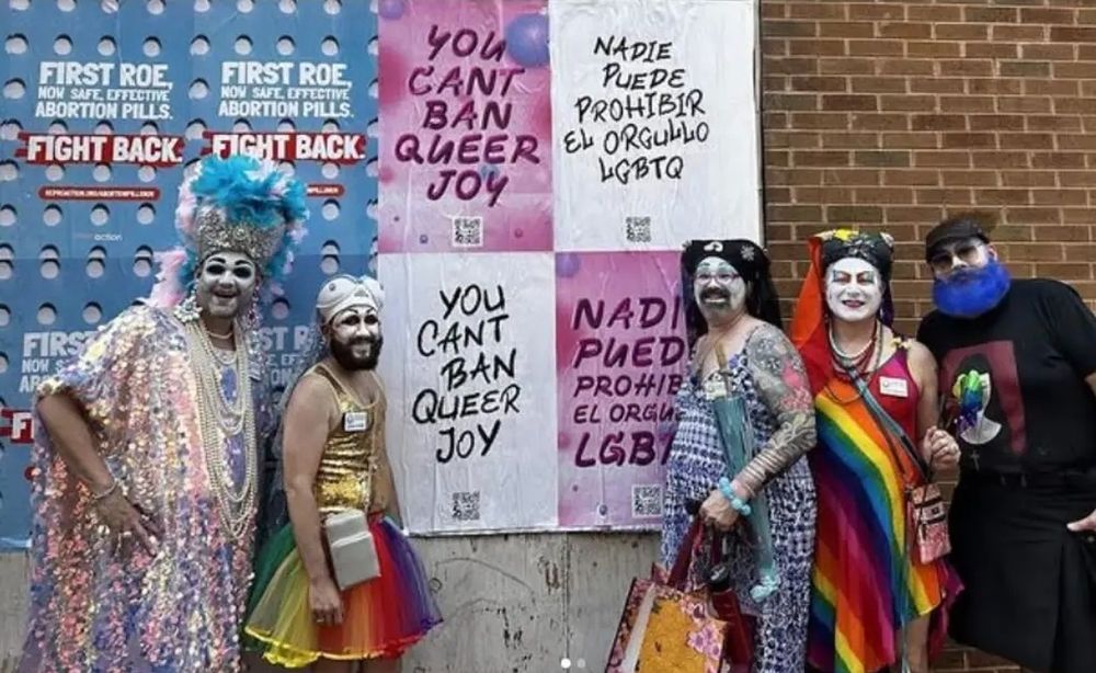 Group of four sisters of perpetual indulgence standing in front of a brick wall that has been plastered with posters that read "You can't ban queer joy."
