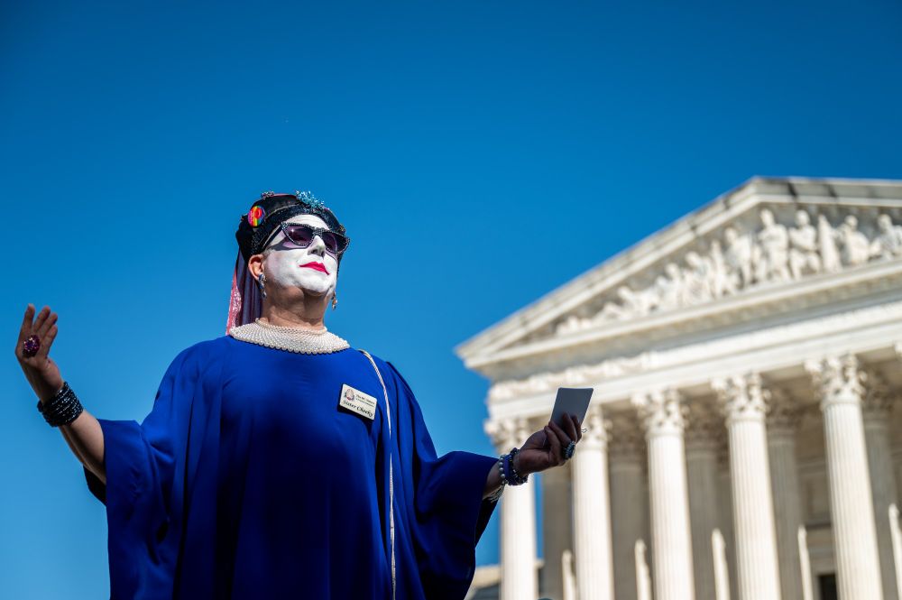 Sister Cheeky dressed in a blue dress with her hands raised out in celebratory prayer, framed against a clear blue sky and the United States Supreme Court building