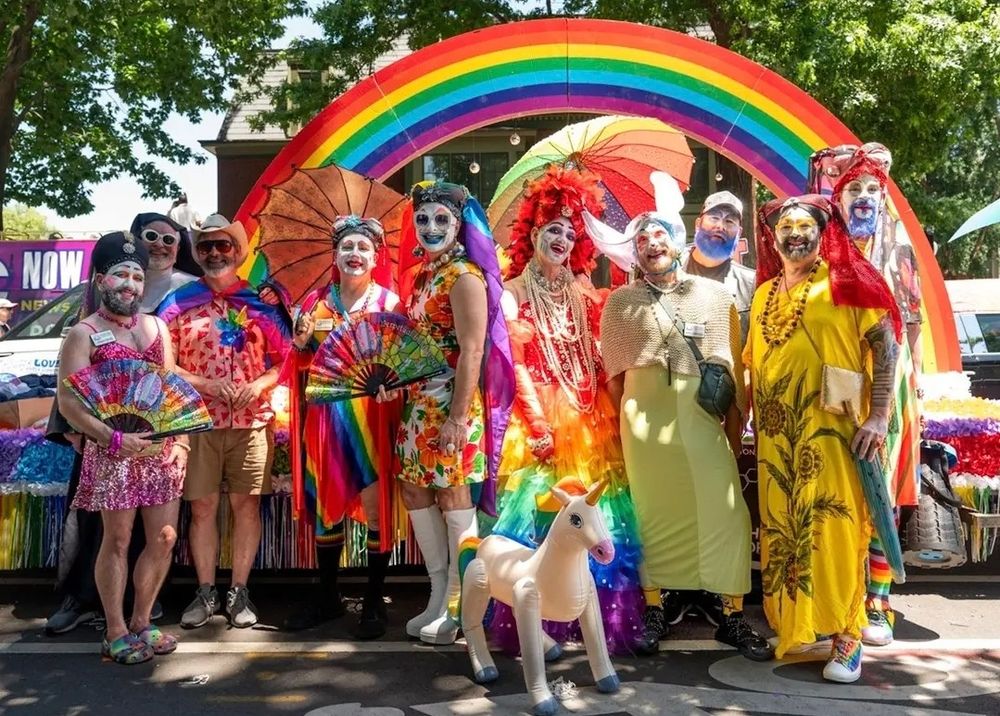 10 Sisters of Perpetual Indulgence dressed in colorful rainbow dresses and fantastic makeup standing in front of a rainbow arch float and a blow up unicorn
