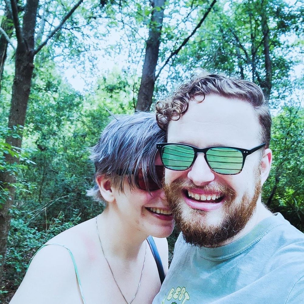 One person has short, gray hair and wears sunglasses, while the other, with curly hair and a beard, wears black-framed sunglasses and a light-colored shirt. They are smiling, with their faces close together