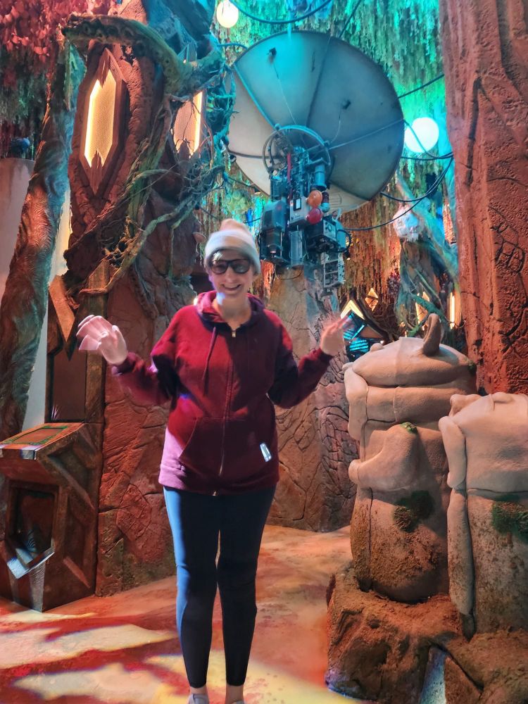 A person stands beside a satellite dish, an artificial tree, and artificial rock formations at Meow Wolf Houston.