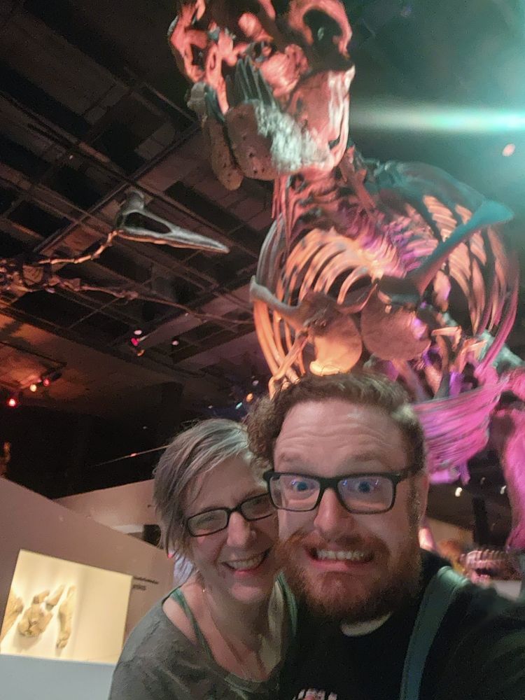 Two people smiling and taking a selfie in front of a large skeleton in a museum.