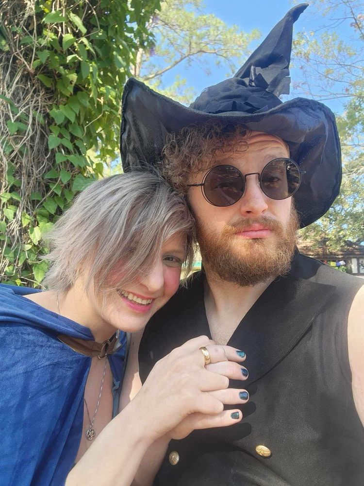 One person with short, grayish hair is smiling and leaning on the other person, who is wearing a large black hat and round sunglasses. This person has curly, reddish hair and a beard, wearing a black vest with gold buttons. Behind them, green leaves are visible under a clear blue sky.