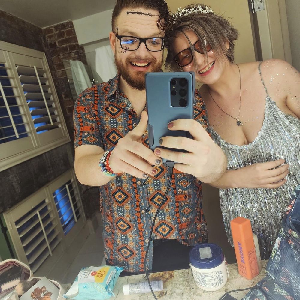 Two Swifties smiling and taking a selfie in a bathroom, standing in front of a mirror with toiletries and personal items on the counter.