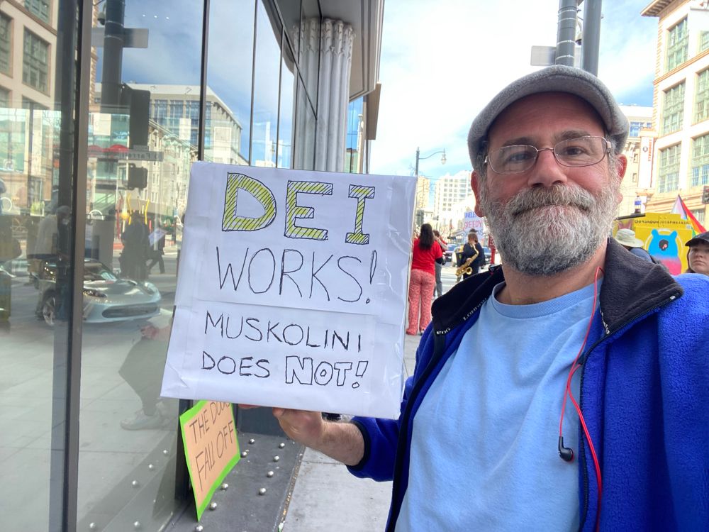 Poster holding a protest sign: “DEI works! Muskolini does not!”