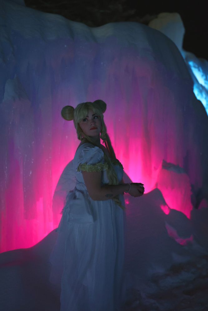 cosplayer dressed as sailor moon in princess serenity form, looking over their shoulder as they stand in front of an ice sculpture lit up in shades of pink. 