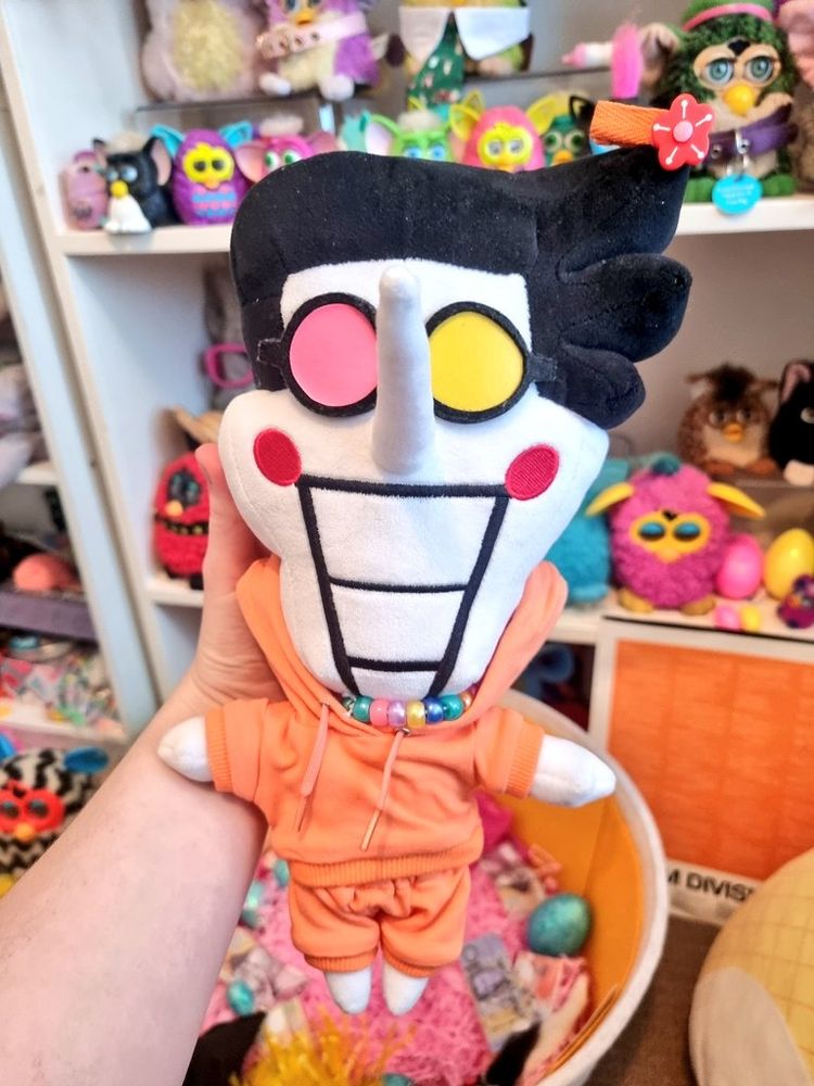 Spamton plush from deltarune wearing a matching set of bright orange hoodie and sweatpants. He has a multicoloured bead necklace. Furby toys are in the background.