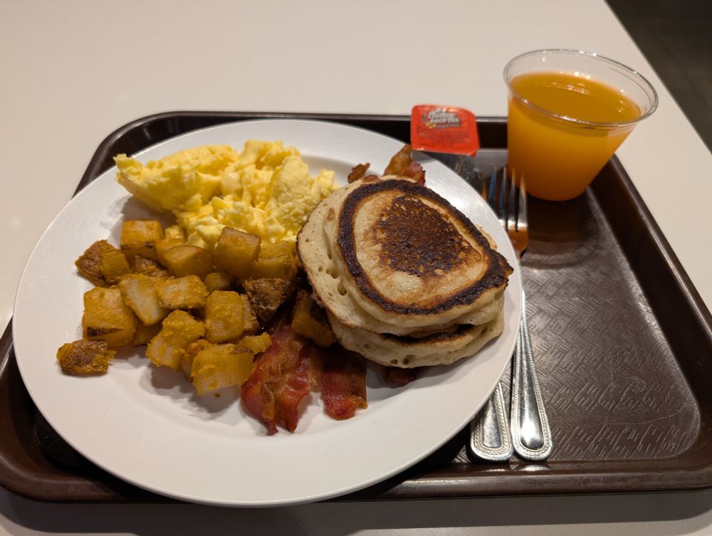 Picture of a plate of scrambled eggs (unseasoned 😞), 4 pieces of bacon, square seasoned potatoes, 2 pancakes, a knife and fork, a cup of juice, and a small rectangle container of Mrs. Butter-worth's syrup. All sits in a brown tray.