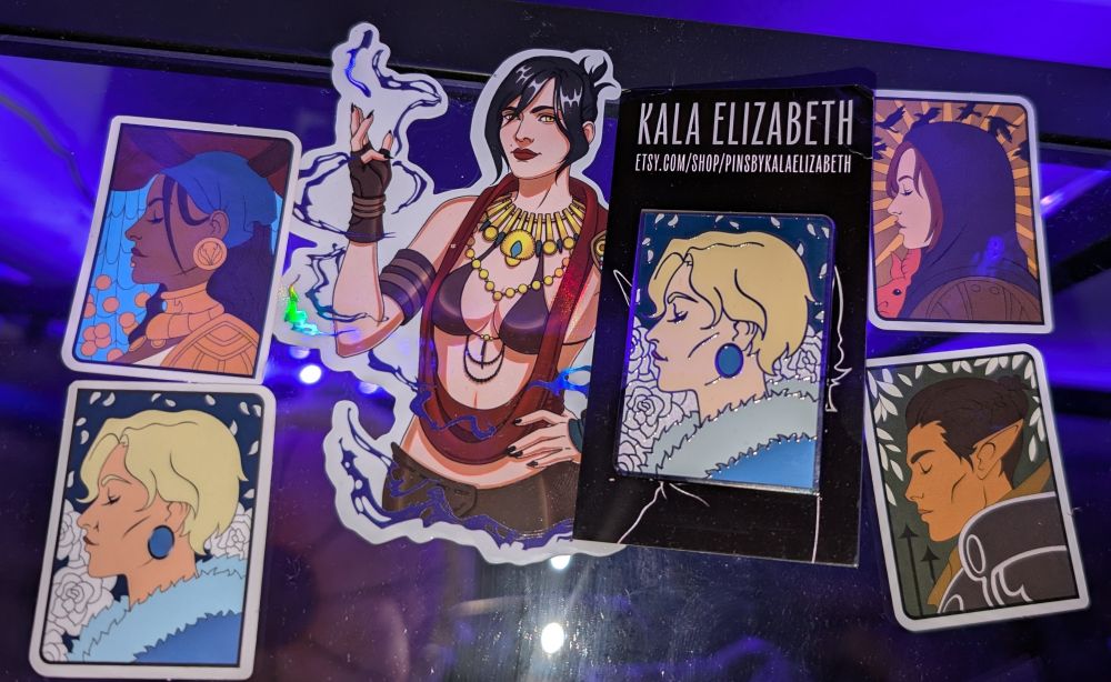 From top left all characters from various Dragon Age games: Isabela left side profile drawing as a sticker,
Maevaris left side profile drawing as a sticker,
A large sticker of Morrigan facing forward with her right hand lifted summoning magic, 
A pin of Maevaris identical to the sticker,
Leliana left side profile drawing as a sticker,
Felassan left side profile drawing as a sticker.