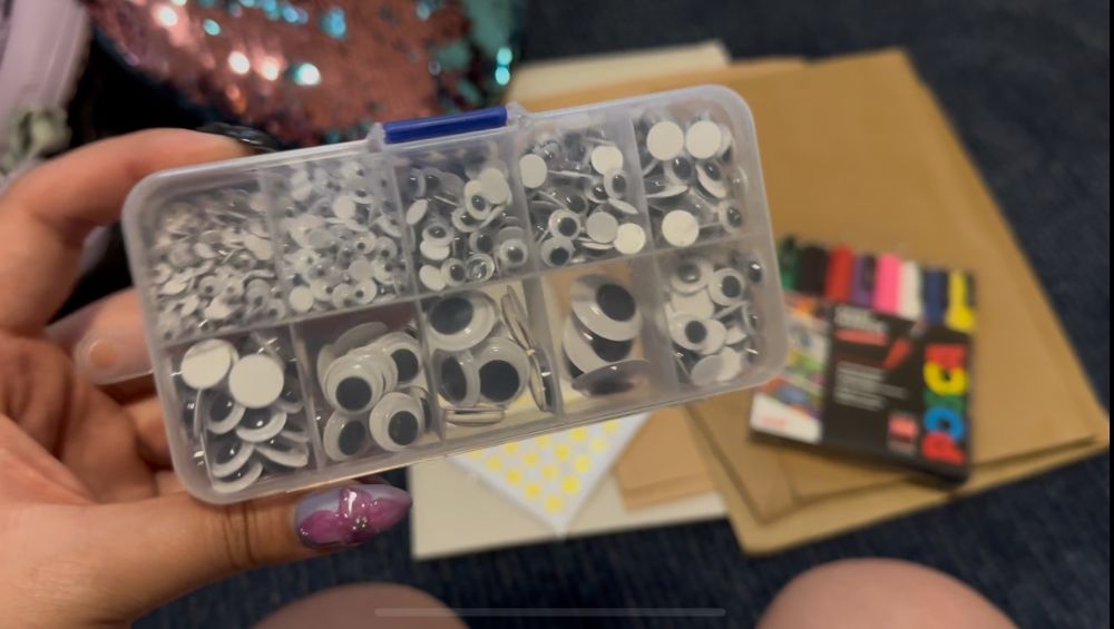 In the foreground, a hand is holding a clear plastic container with ten compartments filled with googly eyes of various sizes. In the blurry background, scattered on the dark blue carpet floor, there’s a sequined pillow, a pack of gold foil star stickers, a white piece of cardboard, brown paper bags, and paint marker pens on top.
