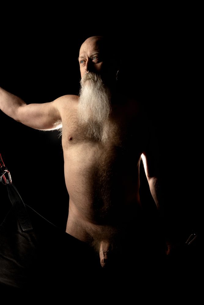 Nude male in his 50's, standing beside in a sling in a darkened room with dramatic lighting. 