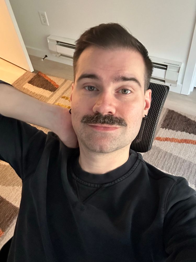 a photo of me holding my phone above me. i’m wearing a black sweatshirt. I have a hand behind my head, and I’m sitting in an office chair with a rug underneath me. 