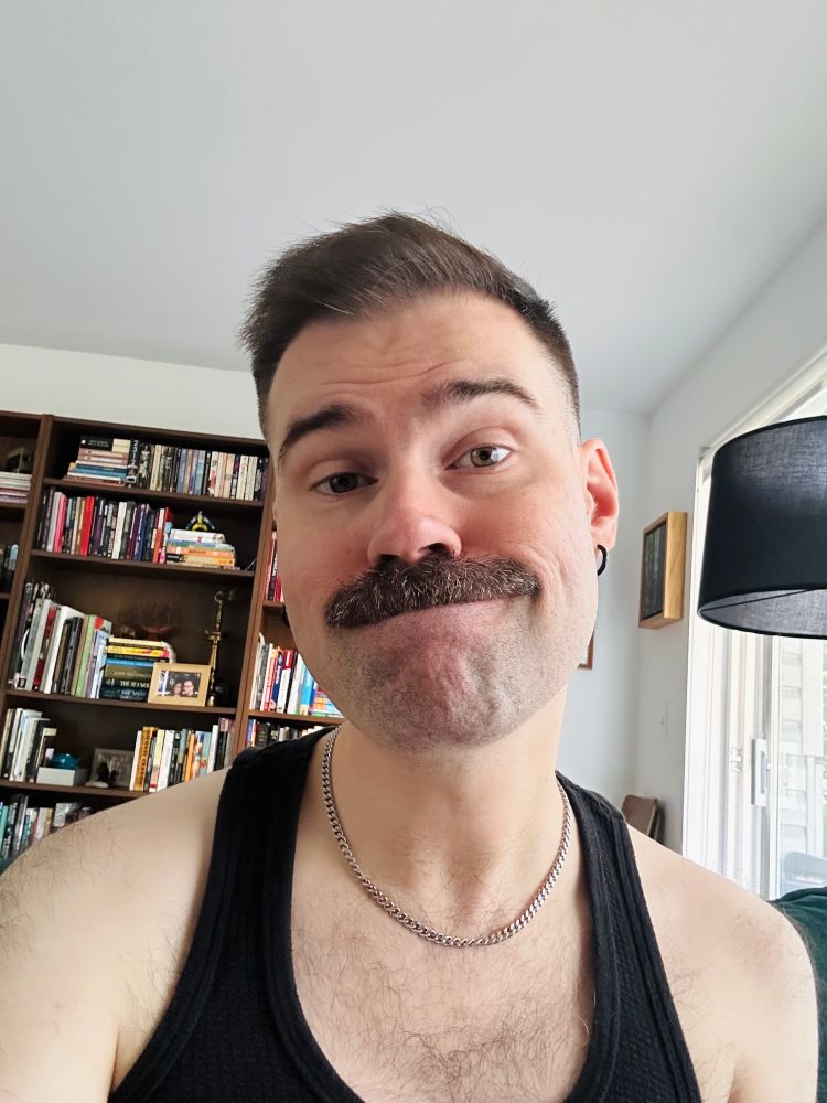 a person with a mustache is wearing black earrings, a chain necklace, and a black tank top. they are sitting in front of a bookshelf.