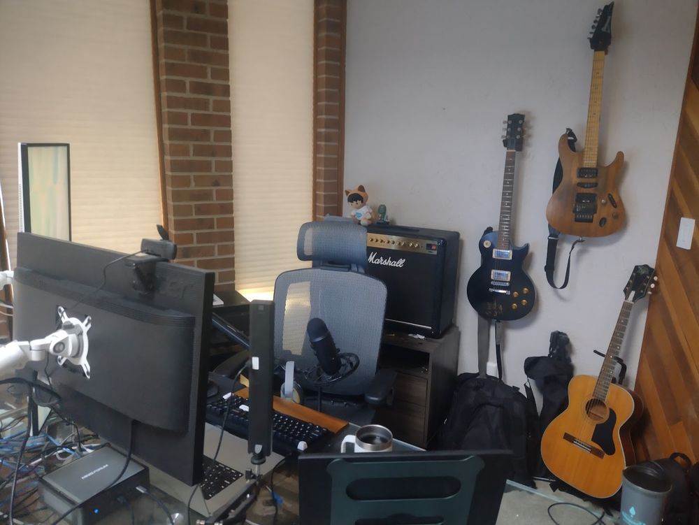 Work from Home setup: behind my computer desk is three guitars: an old regal acoustic, a custom Ibanez S470, and a Gibson Les Paul (autographed by Les Paul). There is also a Marshall DSL40 amplifier, with a little plushy from marketing at work sitting on top

The walls themselves are also interesting. Three walls are shown, one made of drywall, with brick and glass on one side, and wood paneling on the other

Its also kind of generally messy