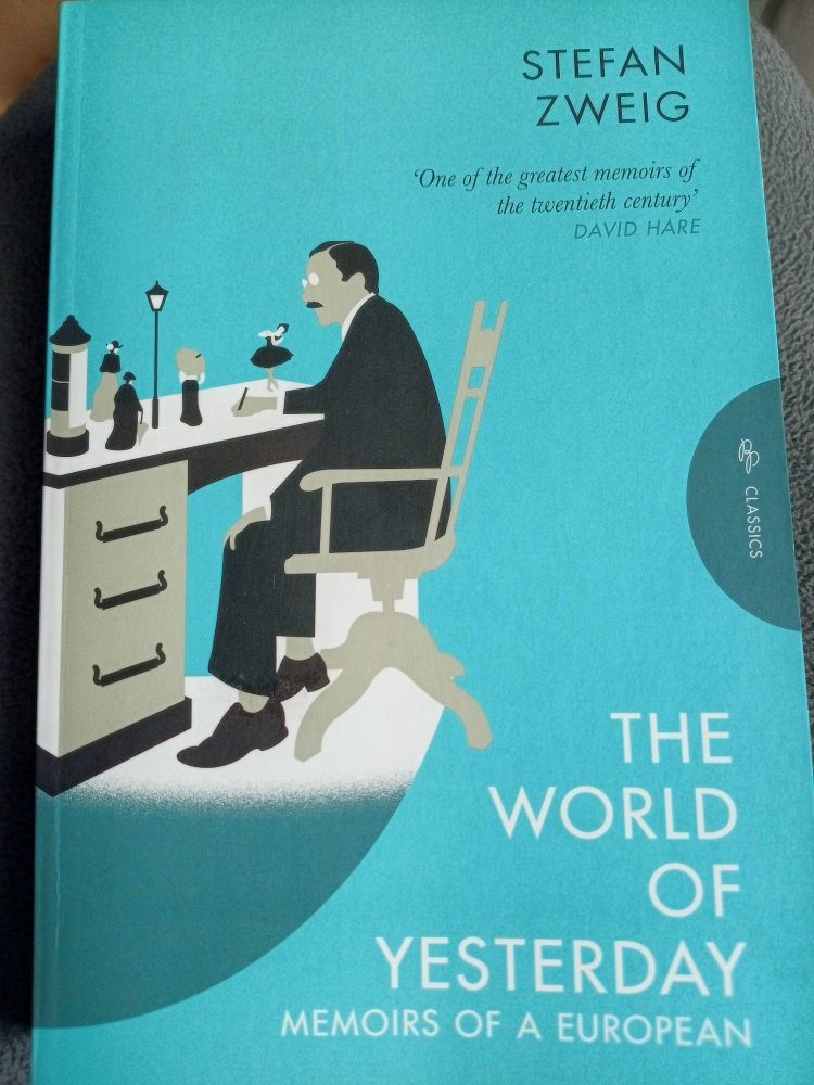 Cover of the book with an illustration of a moustached man sitting at a desk with pen in hand. On the desk is a miniature street scene with a lamppost, an advertising column, a ballerina and three women walking along in typical turn of the century dress - hats, long skirts - two of them are holding umbrellas.