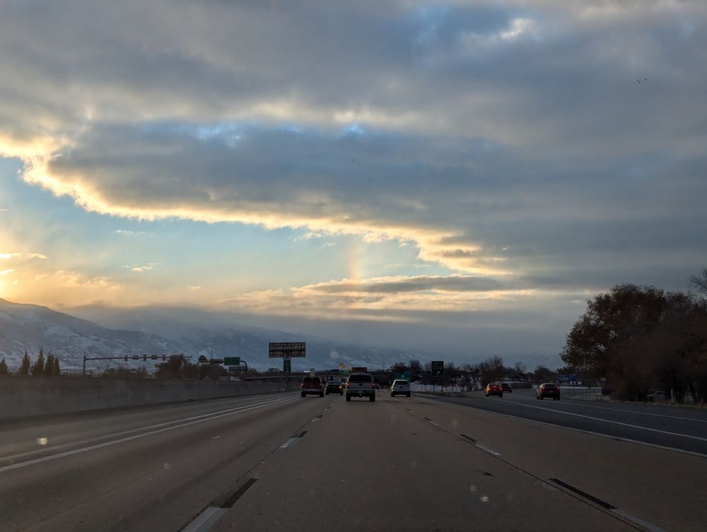 A partly cloudy sky over a freeway with the sun rising above the mountains on the left side of the picture creating a rainbow stretching vertically above the freeway into the clouds.