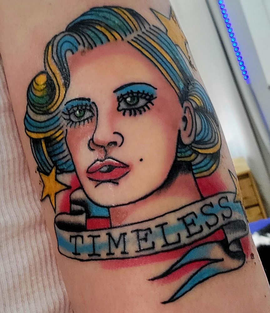 A tattoo of Timeless Toni Storm's face done in color, with her blonde hair represented by light blue and gold. She has blue eyeshadow and red lipstick. There are three yellow stars around her and a banner below her that reads "TIMELESS" 