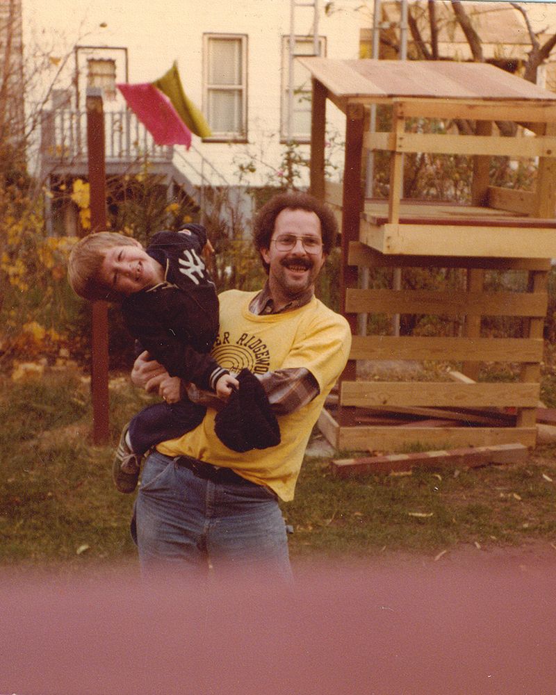 A smiling dad holds his 3 year old son in a Ridgewood Queens backyard.