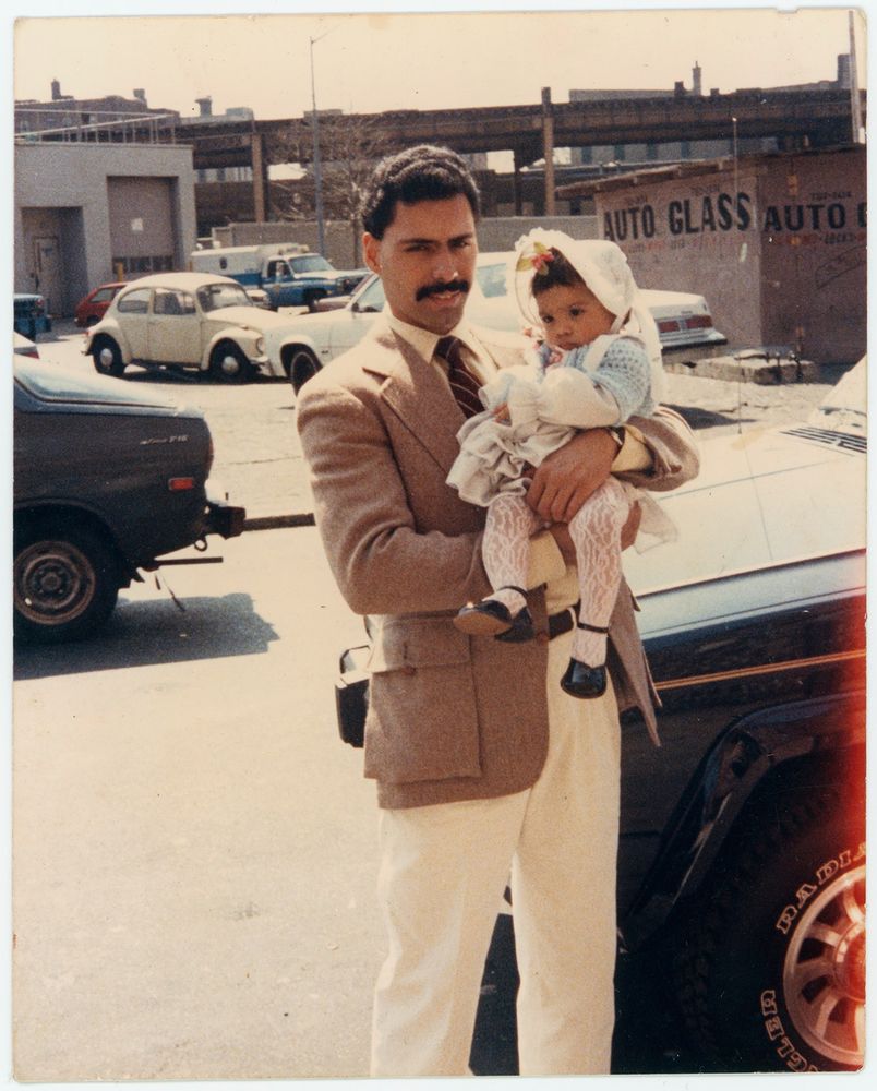 A father in a jacket and tie holds his 1 year old daughter in is arms in front of a parking lot with the elevated tracks for the subway in the background.