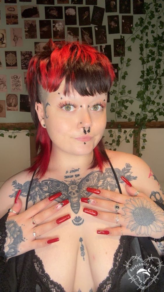 Selfie of Bug. They are wearing a black silk babydoll and robe, and no makeup. They have a red and black mullet, and their green eyes stand out with their red and black lashes. They have their hands resting on their chest showing off their red nails. They are looking at the camera with a small smile. 