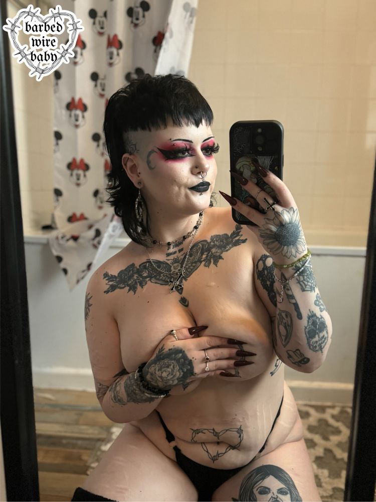 Bug is knelt on the floor wearing a black faux leather thong and thigh high socks, taking a selfie in the mirror. Their hand is covering their nipples, showing off their long stiletto. They have goth makeup on.