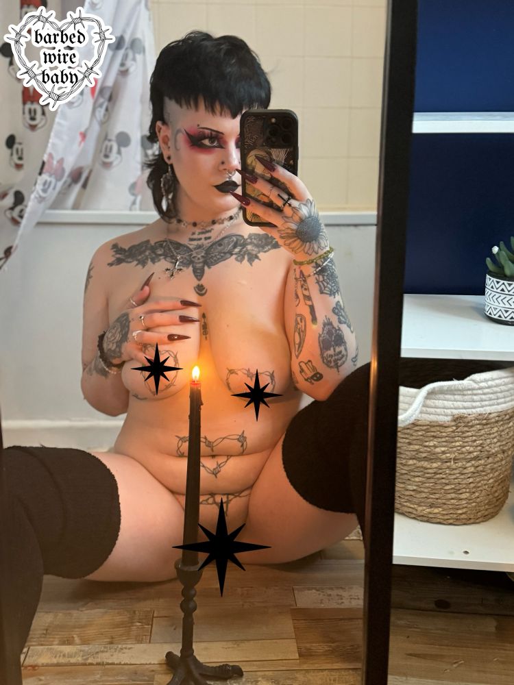 Bug is sat with their legs, nude, in front of a mirror. One hand is resting on their chest, the other holding their phone. There is a candle in front of them. They have goth makeup on.