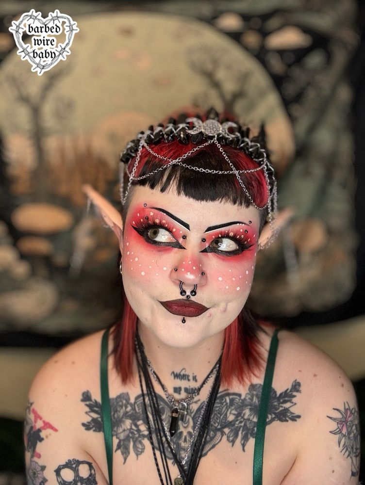 Selfie of Bug who is wearing mushroom inspired makeup. They have elf ears and a crystal headpiece. Behind them is a mushroom backdrop. They are smiling and looking to the side. 

