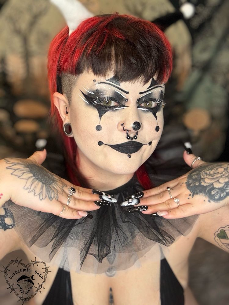 Selfie of Bug. They have black and white clown makeup on, and black and white clown themed nails. They are wearing a black micro bikini, a black and white jester headband, and a black and white tulle clown collar. Their hands are underneath their chin and they are looking at the camera smiling.