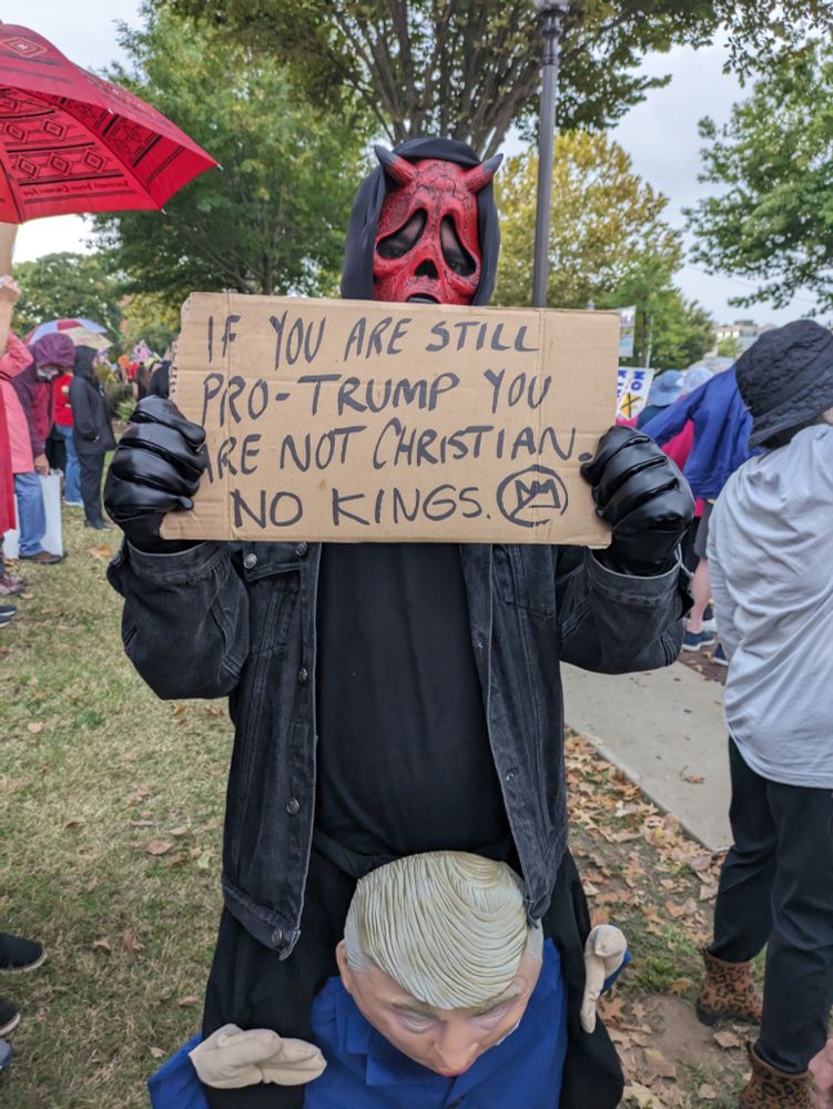 Guy in a costume of a devil riding Trump. Guy is holding a sign that says "If you are still pro-Trump you are not Christian. No Kings."