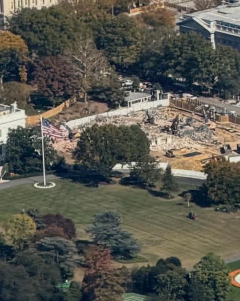 Closer aerial shot of the White House grounds showing the demolished East Wing area. Rubble, construction machinery, and the American flag are visible, marking the site where part of the White House once stood. The image captures the devastation and magnitude of the loss.

