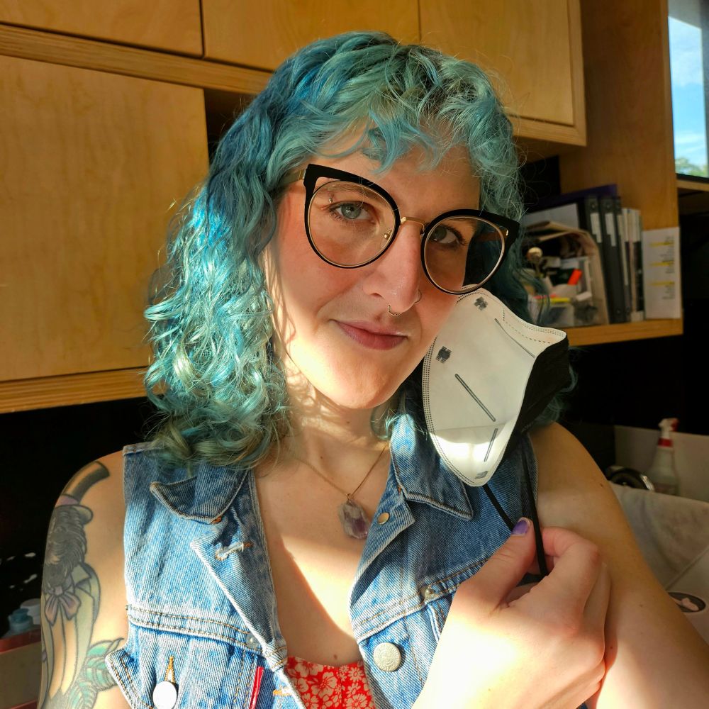 lyra pulling aside her mask. her blue hair is down and she is wearing cat eye glasses, and amethyst necklace, a red floral dress, and a blue denim vest