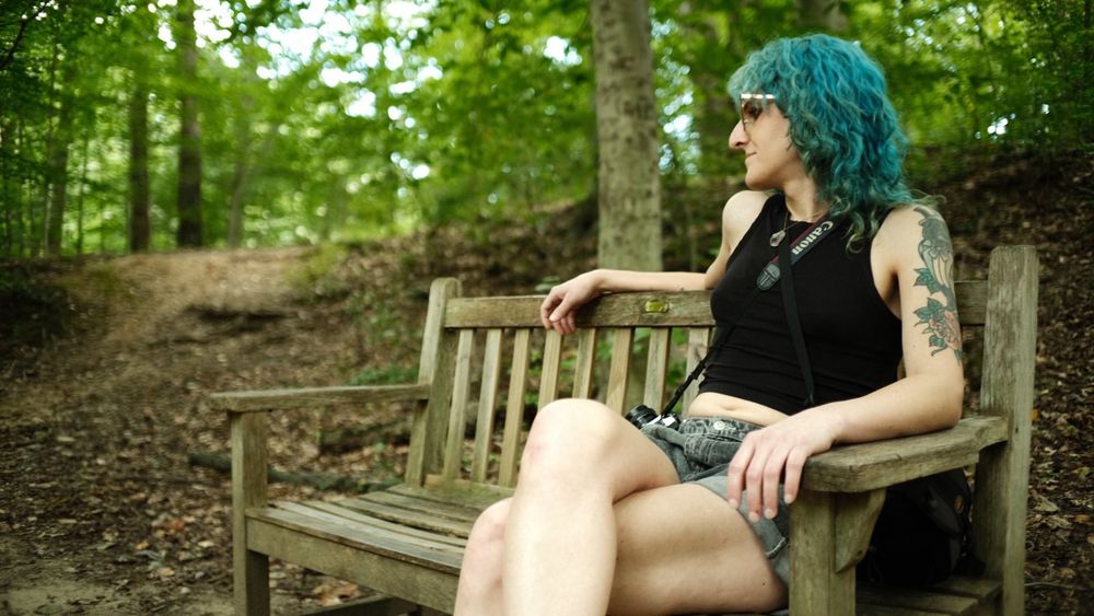 lyra sits on a bench in the woods. she is on the left side of the bench and resting her right arm on the back of the bench while she looks down and to her right.