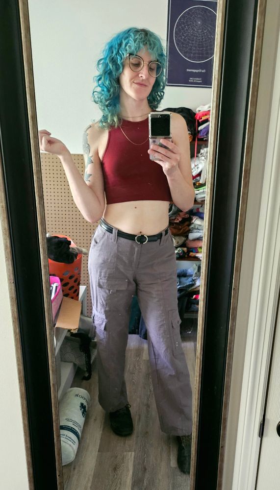 lyra in a couple delicate chain necklaces, a cropped red tank top, grey cargo pants, and black doc martens in her mirror