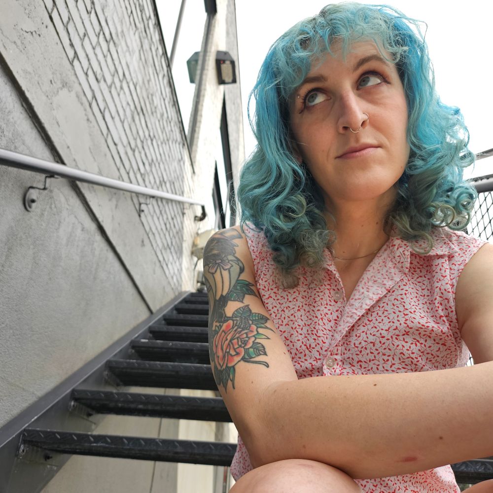 lyra with her blue loose curls down, wearing a white and red leaf pattern sleeveless blouse. she's sitting on a metal staircase outside