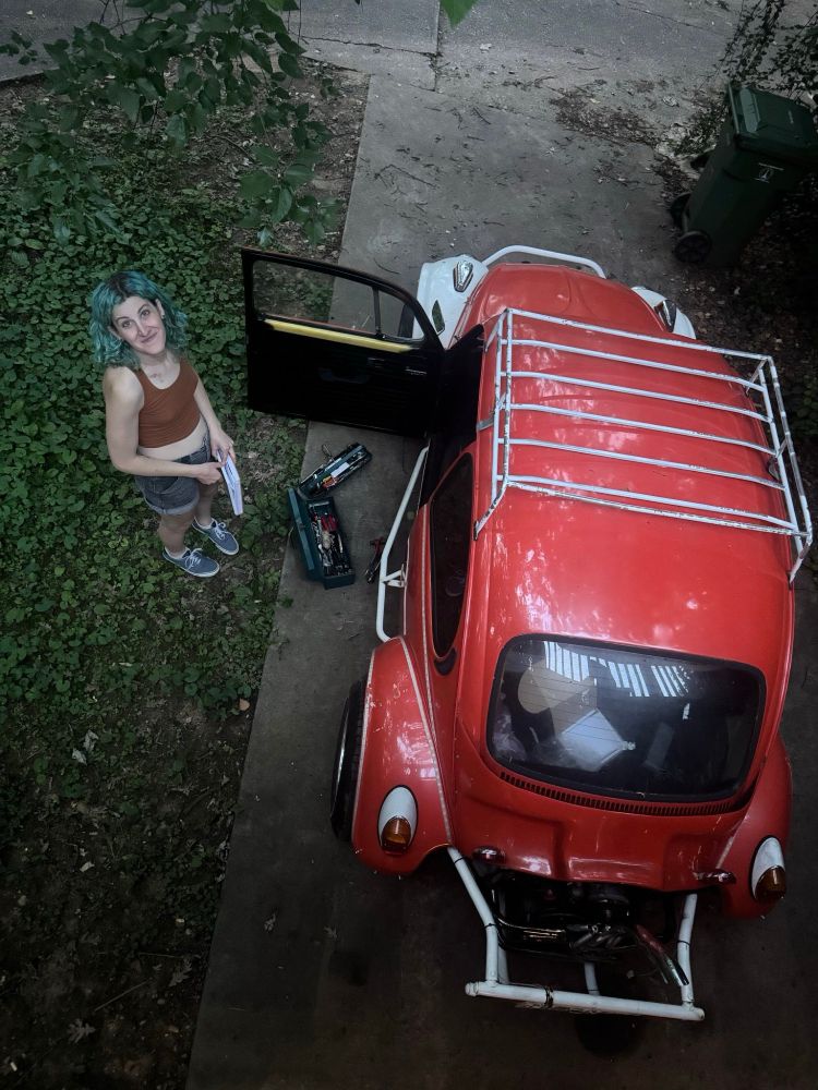 shot from above, lyra is looking up while holding a manual. she is next to an orange vintage vw baja bug with white front fenders and a white roof rack. the vw is parked in a driveway