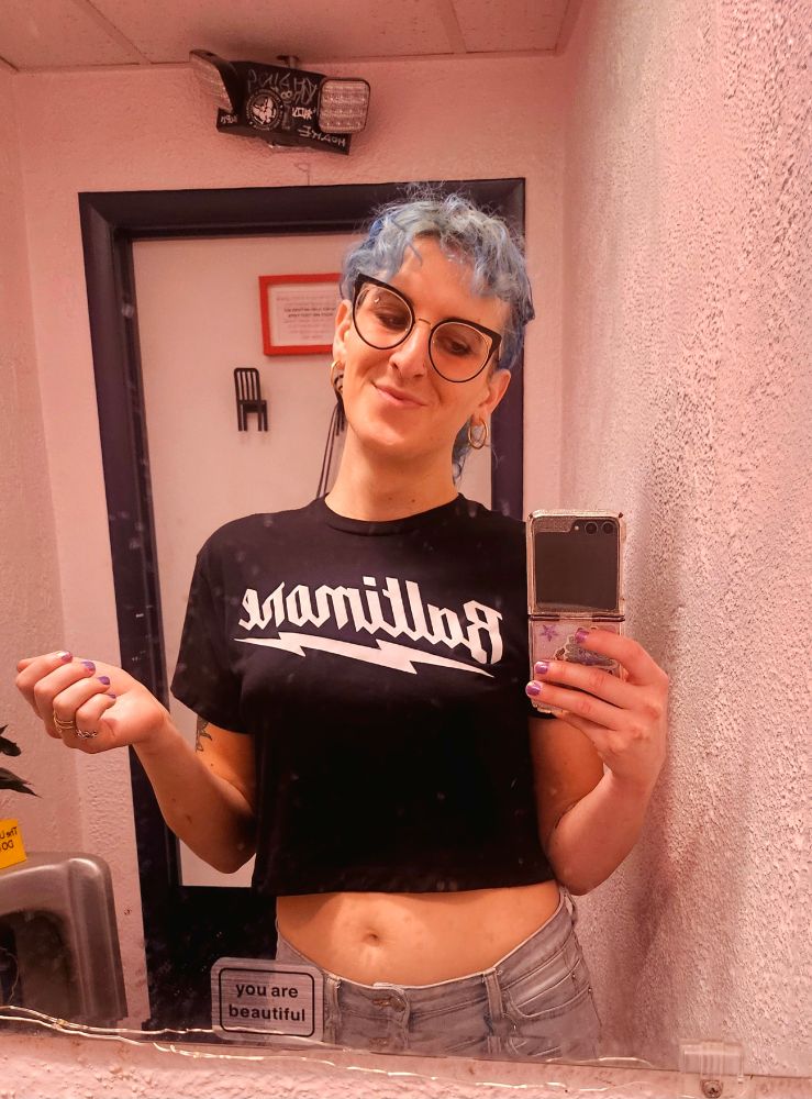 lyra giving a tiny scrunchy smile in a bathroom mirror. there is a sticker on the mirror that says "you are beautiful". lyra is wearing a cropped black t-shirt that says "Baltimore" with a lightning bolt underneath it. she's holding her gay lil gameboy advance sp-looking flip phone