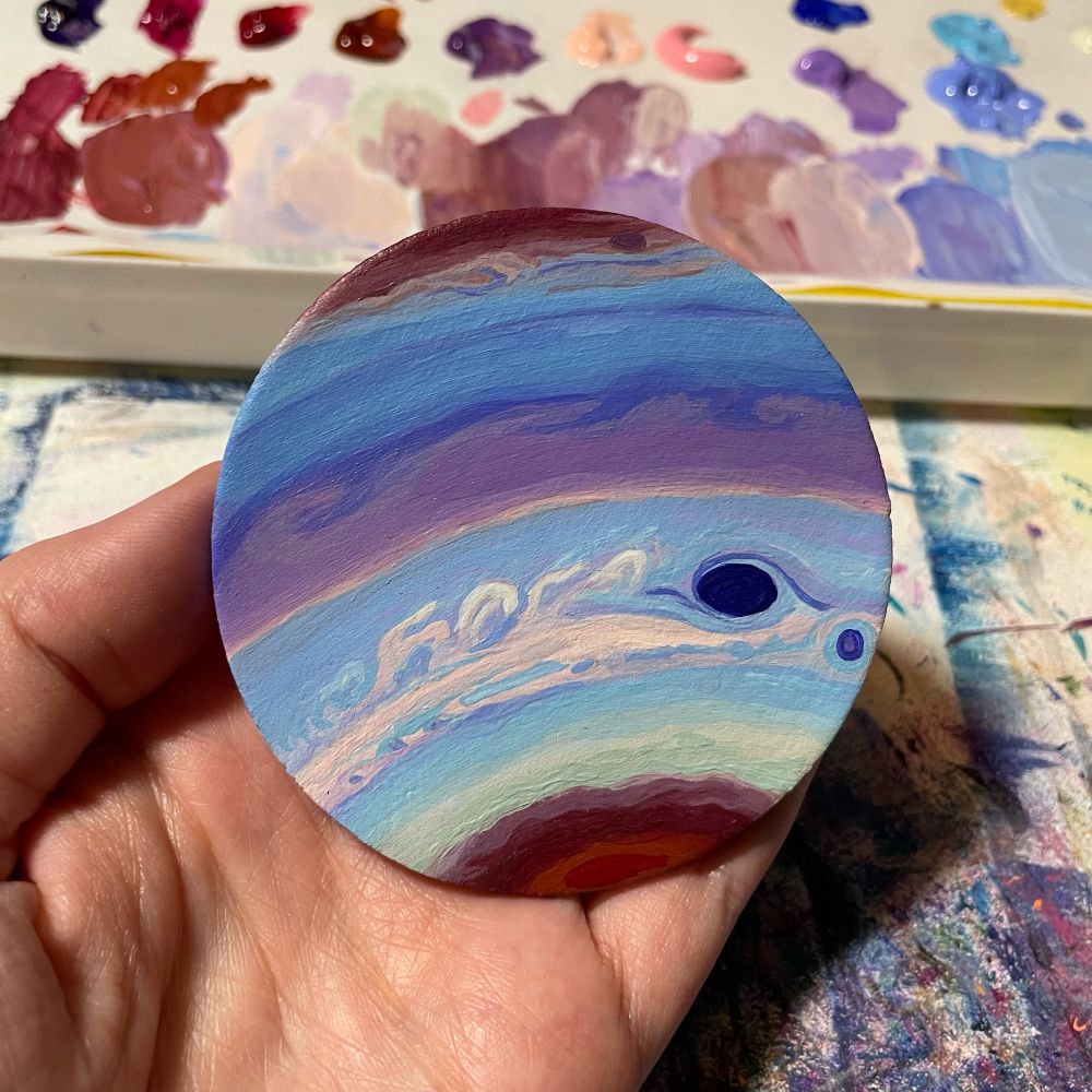 Small round clay piece painted to look like Jupiter in false color. The Great Red Spot is dark purple. The poles are deep red and magenta. The cloud bands are purple and light blue.