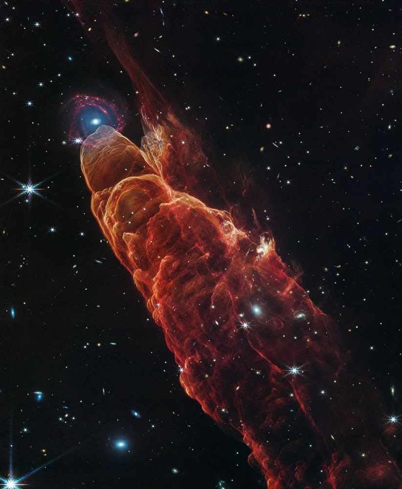 Black background filled with stars and distant galaxies. From the bottom right to the top left is a red and orange pillar. Ripples move through the material like the wake or a boat. At the tip of the pillar happens to be a bright blue galaxy core with deep red spiral arms. 