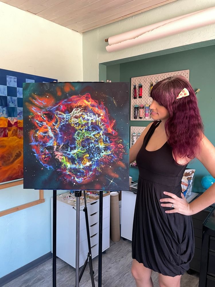 Me in a black dress with a pizza clip in my hair standing next to an easel holding a 24x24” oil painting. The painting is a complex explosion of color in a roughly circular shape with bright oranges, pinks, purples, blues, greens, and yellow. The outside of the circle is a deep teal with peach colored stars. There are 3 subtle triangles that single out a certain wavelength range, and are thus slightly different colors than the surroundings. 