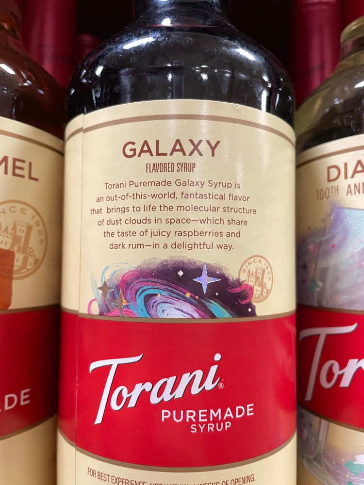 Back of the bottle which reads: Torani Puremade Galaxy Syrup is an out-of-this-world, fantastical flavor that brings to life the molecular structure of dust clouds in space-which share the taste of juicy raspberries and dark rum—in a delightful way.
