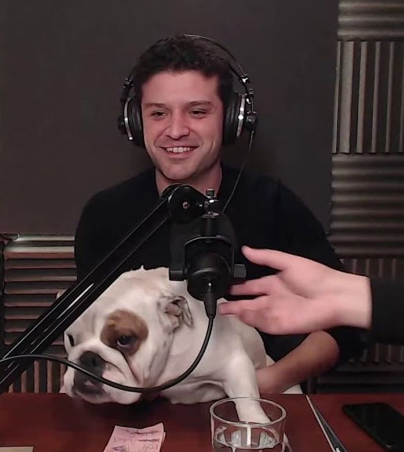 Foto del cómico Miguel Maldonado, tras un micro y llevando auriculares. Está sentado tras una mesa, y en su regazo hay una perra bulldog inglés, blanca con una mancha marrón en el ojo derecho 