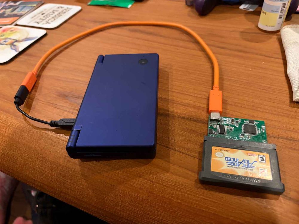 Photograph of a blue-purple Nintendo DSi with a black adapter plugged into it, and into the adapter an orange USB-C cable which is connected to a Game Boy cart reader which has a GBA cartridge plugged in.