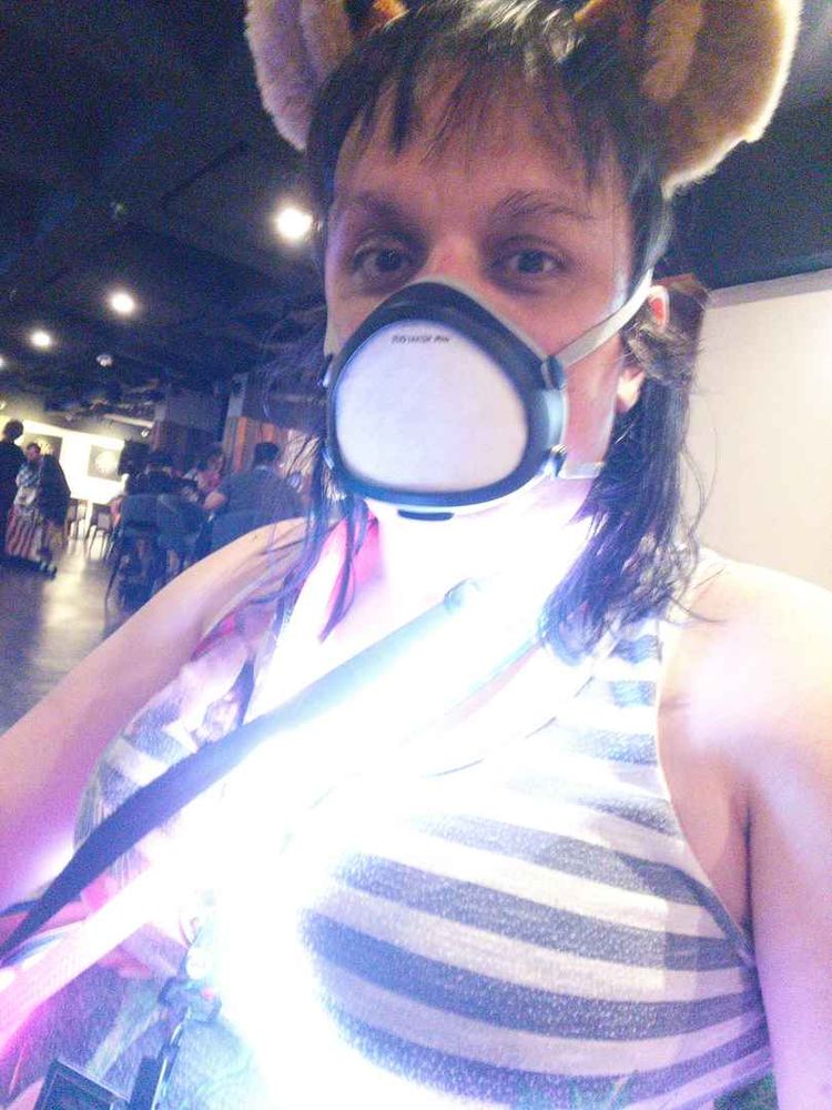 A white girl in cat ears and a black-rimmed flo mask is glowing with lights around her shoulders in a very grainy picture