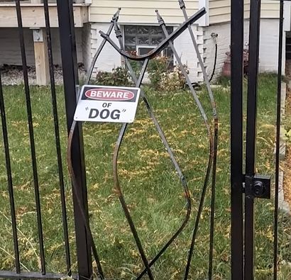 An iron gate which has been mangled, bearing a plastic sign which reads, "BEWARE OF 'DOG'".