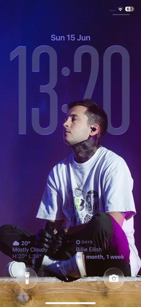 iPhone wallpaper on iOS 26. Picture of Tyler Joseph sat on stage with a purple background, the time has a glass effect and extends behind him